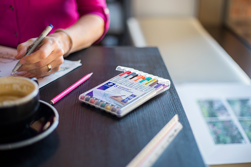 Close up shot of a woman drawing with coloured pens, a ruler and cup of coffee on the table